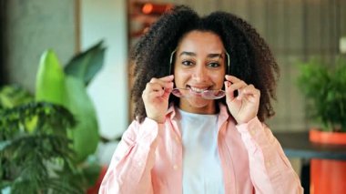 Neşeli genç Afrikalı Amerikalı kadın bilişim uzmanı gündelik pembe tişört beyaz tişört şık gözlük takıyor. Kendinden emin bir şekilde gülümsüyor, canlı yeşil bitkilerin arasında poz veriyor, pozitif duygular.