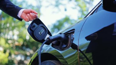 Adamın biri şarj aletini arabadan çıkardı. Dışarıdaki siyah elektrikli araçtan şarj kablosunu çıkarıyor. Sahne, çevre dostu taşımacılığa ve sürdürülebilir enerjiye bağlılığı resmediyor.