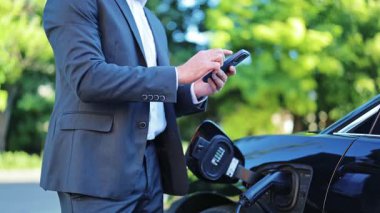 Koyu renk takım elbiseli profesyonel bir işadamı, sakin ve odaklanmış hisseder, siyah elektrikli arabası dışarıda şarj olurken akıllı telefonunu kullanır. Adam arabanın yanında duruyor, modern ulaşım çözümleri..