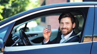 Takım elbiseli yakışıklı işadamı elinde modern araba anahtarlarıyla yeni siyah arabanın içinde gururla gülümsüyor. Profesyonel bir adam heyecanlı, mutlu, başarılı ve araç alımından emin görünüyor..