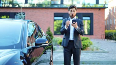 Başarılı bir iş adamı, koyu takım elbiseli, cep telefonuyla zaferi kutluyor. Modern bir ofisin dışında siyah lüks bir arabanın yanında duruyor. Heyecanlı ve muzaffer hissediyor..