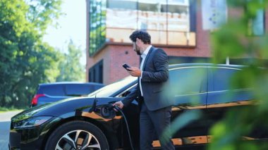 Zeki takım elbiseli başarılı bir işadamı açık havada parlak siyah elektrikli araç şarj ediyor. Akıllı telefon ile meşgul, memnuniyet gösteren ve modern, çevre dostu ulaşım çözümlerini kucaklayan.