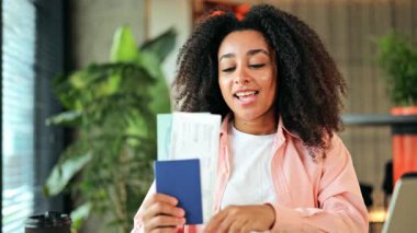 Pembe tişört ve beyaz tişört giyen heyecanlı Afro-Amerikalı kadın, evde otururken elinde bir pasaport ve uçak bileti tutuyor, harika bir yolculuk bekliyor..