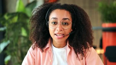 Kıvırcık saçlı ve gözlüklü neşeli Afro-Amerikan kadın profesyonel, pembe gömlek ve kulaklık giyiyor, modern bir ofiste çevrimiçi video görüşmesinde güvenle gülümsüyor..