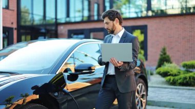 Takım elbiseli odaklanmış iş adamı dışarıda dizüstü bilgisayarda çalışıyor. Şık elektrikli arabası modern bir istasyonda, sürdürülebilir taşımacılığa olan bağlılığını gösteriyor. Sakin görünüyor..