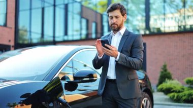 Açık havada duran şık takım elbiseli profesyonel bir işadamı modern elektrikli bir arabanın yanında akıllı telefonuyla neşeli bir şekilde meşgul oluyor. Bu içerikli erkek yeşil teknolojiyi kucaklıyor..