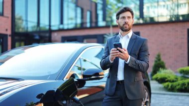 Parlak takım elbiseli mutlu bir iş adamı, açık hava istasyonunda şarj olan modern bir elektrikli aracın yanında akıllı telefonuyla gülüyor, sürdürülebilir teknoloji evlat edinme sergiliyor..