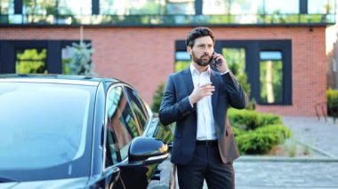 Kendine güvenen, koyu takım elbiseli profesyonel iş adamı, lüks siyah arabanın yanında dururken akıllı telefonuyla konuşuyor. Uzman, çağdaş tuğla bina yakınlarında dışarıda olmaktan memnun görünüyor..
