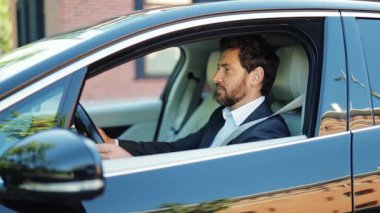 Şık bir takım elbise giymiş, modern lüks arabasında oturan başarılı işadamı, kentli bir sokakta kendinden emin, mutlu bir gülümsemeyle kameraya bakıyor..