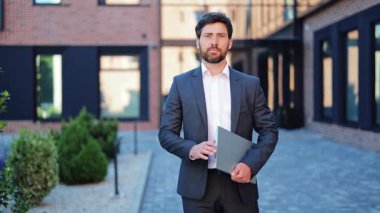 Profesyonel işadamı, şık bir takım elbise giymiş, dışarıda dizüstü bilgisayarla duruyor, modern bir ofis binasının yakınında mutlu bir şekilde gülümsüyor, başarıyı ve yönetici varlığını temsil ediyor..