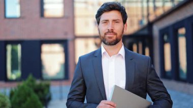 Zeki gri takım elbiseli ve beyaz gömlekli profesyonel işadamı şehirde neşeli bir şekilde gülümsüyor. Kendine güvenen erkek dizüstü bilgisayar tutuyor, başarıyı, başarıyı ve olumlu kariyer hedeflerini temsil ediyor..