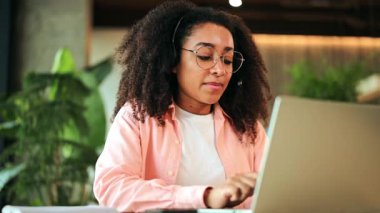 Kulaklık takan neşeli Afrikalı Amerikalı kadın modern bir ofiste dizüstü bilgisayar kullanıyor. Pembe bir gömlek giyip, mutlu ve üretken bir şekilde profesyonel internet iletişimi kuruyor..