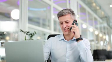 Açık mavi gömlekli kendine güvenen işadamı cep telefonuyla konuşuyor, modern bir ofiste dizüstü bilgisayarla çalışıyor, etkili bir iletişim ve konsantrasyon gösteriyor..