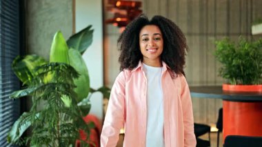 Pembe tişörtlü, beyaz tişörtlü, gülümseyen ve onaylayan neşeli Afro-Amerikan kadın. Modern ofis ortamında memnuniyetini ve onayını ifade ediyor, mutlu hissediyor..