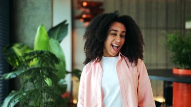 Kıvırcık saçlı güzel bir Afro-Amerikalı kadın, beyaz bir tişörtün üzerine günlük pembe bir gömlek giyiyor, parlak bir iç mekanda neşe içinde göz kırpıyor ve gülümsüyor. Mutluluğu ve güveni ifade eder.