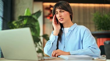 Stresli profesyonel iş kadını, ofis masasında dizüstü bilgisayarı üzerinde çalışırken acı verici bir baş ağrısı ve göz yorgunluğu yaşıyor. Rahatsızlık ve yorgunluk hisleri çok açık..