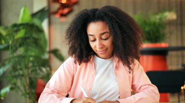 Pembe tişörtlü güzel Afro-Amerikalı kadın bir kafede stilisyonla çalışıyor. Dikkatlice odaklanıyor, sonra mutlu bir şekilde gülümsüyor, neşe ve yaratıcılığını ifade ediyor..