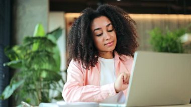 Profesyonel Afro-Amerikan iş kadını, pembe gömlekli, laptopunda özenle çalışıyor, parlak bir ofiste mutlu bir şekilde gülümsüyor. Mutlu ve ilham dolu hissediyor..