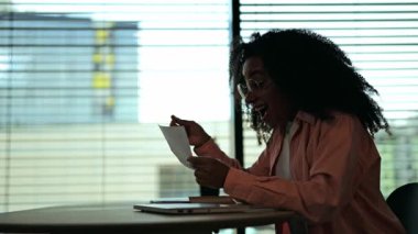 Rahat bir gömlek giymiş heyecanlı bir Afrikalı Amerikalı kadın, parlak, modern bir ofiste belge okumaktan zevk alıyor. Neşeli tepkisi sürpriz ve başarıyı gösteriyor..