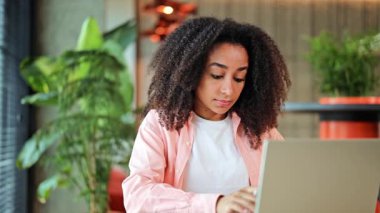 Pembe tişörtlü ve beyaz tişörtlü profesyonel Afro-Amerikan iş kadını özenle bir dizüstü bilgisayar üzerinde çalışıyor, parlak, modern ofis alanına notlar alıyor, konsantrasyon ve verimliliği gösteriyor..