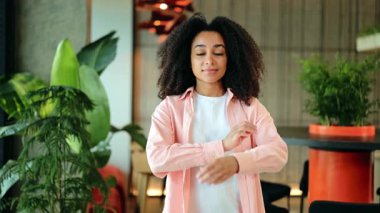 Pembe tişörtlü, beyaz tişörtlü, kendine güvenen Afro-Amerikan kadın modern bir ofiste kollarını kavuşturarak gülümsüyor. Modern İşyerinde Gülümseyen Afrikalı Amerikalı Kadın Portresi.