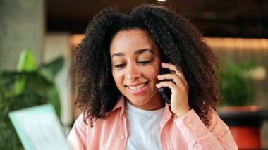 Pembe tişörtlü heyecanlı Afro-Amerikalı kadın elinde pasaport ve biletlerle telefonuyla hareketli bir şekilde konuşuyor. İçeride neşeyle ışıldıyor, yaklaşan seyahat macerasına hazır..