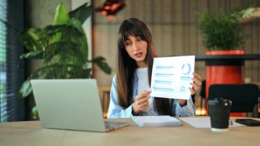 Açık mavi gömlekli bir iş kadını, modern bir ofisten internet üzerinden veri tablolarını güvenle açıklayan bir toplantı düzenliyor. Hayati bilgiler sunuyor ve ciddi, odaklanmış bir duygu aktarıyor..