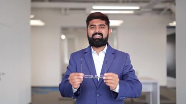 Mavi takım elbiseli ve beyaz gömlekli profesyonel Hintli işadamı neşeyle gülümsüyor. Kendine güvenen erkek model, modern bir ofiste gözlük takıyor, kollarını kavuşturuyor..