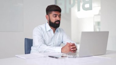 Başarılı Hintli işadamı, parlak ve modern bir ofiste çevrimiçi bir video görüşmesine aktif olarak katılıyor. Açık mavi gömlekli profesyonel bir adam meşgul ve odaklanmış görünüyor..