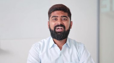 Açık tişörtlü neşeli Hintli işadamı modern bir ofiste güvenle gülümsüyor. Mesleki davranışları olumlu duyguları ve başarıyı yansıtıyor..