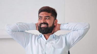 Açık mavi tişörtlü neşeli Hintli profesyonel adam gülümsüyor, elleri başının arkasında, parlak bir ofis ortamında dinleniyor. Memnun iş adamı kendini huzurlu hissediyor.