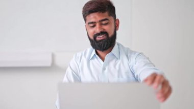 Neşeli Hintli işadamı, açık tişört giymiş, bilgisayarını parlak ofiste kapatıyor, başarılı bir günün ardından kendini rahat ve tatmin olmuş hissediyor. Hintli erkek huzur ve refahın tadını çıkarıyor..