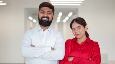 Kendine güvenen Hintli iş adamı ve zarif Hintli iş kadını, şık giyinmiş, parlak bir ofiste kollarını kavuşturarak gülümsüyor. Mutluluğu ve başarıyı ifade etmek.