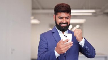 Mavi takım elbiseli heyecanlı Hintli işadamı modern ofisteki akıllı telefonda zafer ve mutluluk dolu haberleri kutluyor. Profesyonel erkek zafere ulaştı..