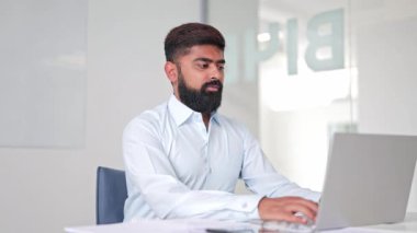 Hasta Hintli işadamı, modern ve parlak bir ofiste dizüstü bilgisayarda çalışır. İş adamı açık mavi gömlek giyer. Duraksamalar, düşünceli ve samimi görünmeler, işi yansıtıyor..