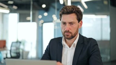 Modern ofisteki düşünceli iş adamı dizüstü bilgisayarla çalışıyor. Odaklanmış, düşünceli, önemli görevlere odaklanmış, takım elbiseli bir uzman. Ciddi bir erkek yatırımcı strateji düşünüyor..