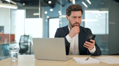 Odaklanmış yönetici işadamı bilgisayarının üzerinde çalışırken, aynı zamanda akıllı telefonunu da kullanıyor, parlak, modern bir ofiste, iş kararlarına odaklanmış ve düşünceli görünüyor..