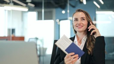 Profesyonel takım elbiseli neşeli bir iş kadını, modern ofiste telefonda konuşuyor, elinde pasaport ve biletler var. Yaklaşan seyahatleri bekliyor, neşeli ve başarılı hissediyor..