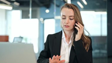Siyah ceketli ve beyaz gömlekli odaklanmış iş kadını modern bir ofiste belgeleri incelerken ciddi bir telefon görüşmesi yapıyor. Odaklanmış ve meşgul görünüyor..