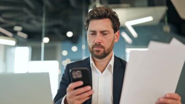 Modern bir ofiste profesyonel bir işadamı, takım elbise giyiyor, belgeleri ve akıllı telefonu dikkatle inceliyor. Odaklanmış konsantrasyonu, düşünceli karar verme mekanizmasını ve kendine güvenen hafif bir gülümsemeyi gösterir..