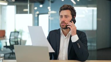 Modern bir ofiste iş belgelerini tartışan resmi takım elbiseli profesyonel işadamı, bir dizüstü bilgisayar üzerinde çalışırken odaklanmış ve meşgul görünüyor..