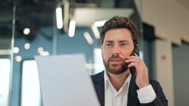 Pozitif iş adamı, takım elbiseli, bir telefon görüşmesi yapıyor. Modern bir ofiste güvenle gülümsemeden önce ciddi bir ifadeyi gösteren belgeleri gözden geçirir.