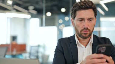 Endişeli bir iş adamı, takım elbise giymiş, modern bir ofiste endişeli görünüyor. Yakışıklı erkek akıllı telefondan üzücü haberler okurken endişeyle ve derin düşüncelerle tepki verir.