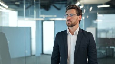 Siyah takım elbiseli ve beyaz gömlekli kendine güvenen bir işadamı gözlüğünü çıkarır ve parlak, modern bir ofiste kameraya neşeyle gülümser. Başarılı ve mutlu görünüyor..