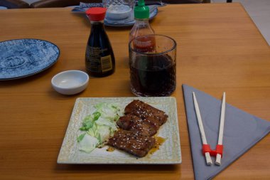 Bir tabak et ve sebze, yemek çubukları ve baharatlarla ahşap bir masada oturur. Sofra kurulumu basit ve zarif, özellikle de yemeğe odaklı.