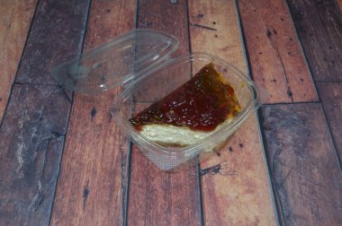 Bir parça cheesecake, ahşap bir masadaki plastik bir konteynerin içinde duruyor. Konteynır açık ve peynirli kek içeride görünüyor. Cheesecake bir dilim çilekli cheesecake gibi görünüyor.