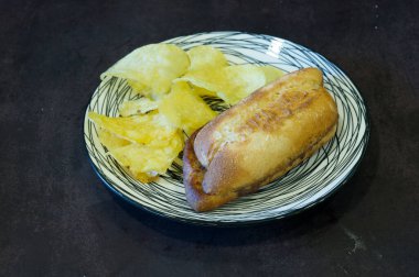 Tabakta bir sandviç ve biraz cips var. Sandviç ikiye bölünmüş ve cipsler de kenarda.