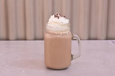 Üzerine krem şanti sürülmüş bir bardak süt. Krem şanti beyaz ve kabarık. Cam bir masanın üzerinde duruyor.