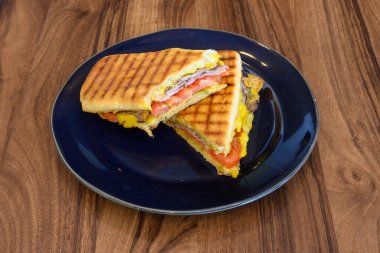 Bir sandviç ikiye bölünür ve mavi bir tabağa yerleştirilir. Sandviç jambon ve peynirle yapılır ve ahşap bir masada servis edilir.