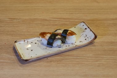 Bir suşi rulosu ahşap bir masada beyaz bir tabakta oturur. Tabak noktalardan ve çizgilerden oluşan bir desenle süslenmiş. Suşi böreği görüntünün ana odak noktasıdır. Lezzetli ve iştah açıcıdır.